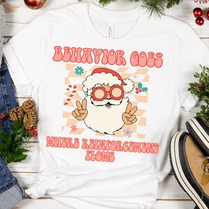 Op de afbeelding: Wit T-shirt met een rode en witte kerstman die een zonnebril draagt en vredestekens maakt. De tekst "Behavior Goes Where Reinforcement Flows" is in roze en wit.