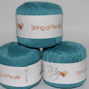 May include: Three teal yarn skeins stacked on top of each other. Each skein has a white label with the text "String of Pearls" and a logo. The yarn has a subtle shimmer, suggesting a blend of fibers.