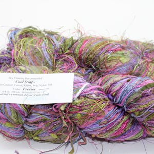 May include: A skein of Cool Stuff yarn in the color Freesia. The yarn is a blend of cotton, rayon, poly, nylon, and silk. The yarn is a mix of green, pink, purple, and silver.