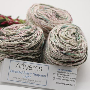 May include: Three skeins of Artyarns Beaded Silk + Sequins Light yarn in a beige, green, and pink colorway. The yarn is 100% silk with glass beads and sequins. The label reads "Artyarns Beaded Silk + Sequins Light 100% Silk w/Glass Beads & Sequins 50g/100m/110yd CLUB GET INSPIRED facebook.com/artya artyarns.com Free Pattern: www.a Enter coupon code: Size 6 US Needles 5"