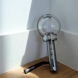 May include: A gray, cylindrical light stick featuring a clear, spherical top dome. The handle displays the name HEESEUNG in iridescent lettering and Korean characters. A black wrist strap with the white text ENHYPEN is attached to the base.