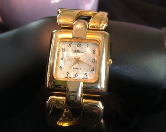 May include: A gold-toned wristwatch with a square face and a linked band. The watch face has black Roman numeral hour markers and a white background with the words "Le Baron" and "Quartz" printed on it.