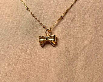 Puede incluir: Un collar de cadena dorada con un pequeño colgante de lazo dorado.