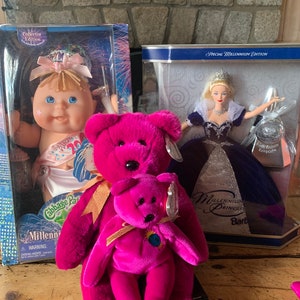 May include: A pink Millennium Princess Beanie Baby bear with a smaller pink bear in its arms. The larger bear has a gold ribbon around its neck. The smaller bear has a gold ribbon around its neck and a blue globe on its chest. The bears are sitting on a wooden surface. There are two boxes in the background, one with a Cabbage Patch Kid doll and the other with a Millennium Princess Barbie doll.