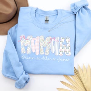 May include: A light blue sweatshirt with the word "MAMA" in a patchwork design. The letters feature floral, gingham, and striped patterns, accented with pink ribbons. Below the word are the names "Oliver", "Ella", and "James". A silver necklace with a star pendant is on the sweatshirt.