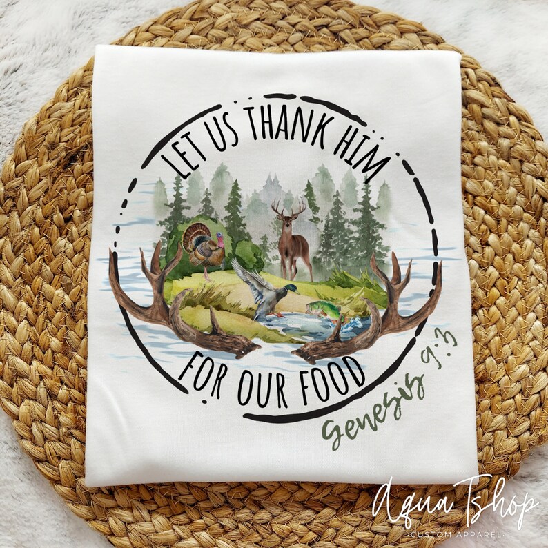 May include: White t-shirt with a black circle design featuring a watercolor illustration of a forest scene with a deer, a turkey, and a duck. The text "LET US THANK HIM FOR OUR FOOD Genesis 9:3" is written around the circle.