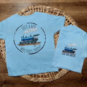 May include: Two light blue t-shirts with a train design. The larger shirt says "Just a boy who loves trains" and the smaller shirt says "Future train conductor".