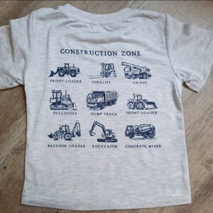 May include: A gray t-shirt with a blue print of construction vehicles. The text "CONSTRUCTION ZONE" is at the top of the shirt. The vehicles are labeled with their names: Front Loader, Forklift, Crane, Bulldozer, Dump Truck, Front Loader, Backhoe Loader, Excavator, and Concrete Mixer.