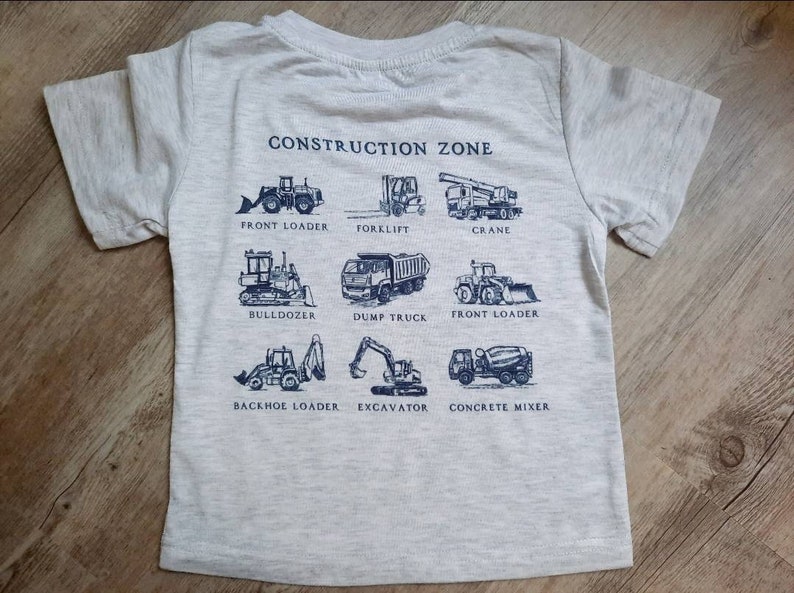 May include: A gray t-shirt with a blue print of construction vehicles. The text "CONSTRUCTION ZONE" is at the top of the shirt. The vehicles are labeled with their names: Front Loader, Forklift, Crane, Bulldozer, Dump Truck, Front Loader, Backhoe Loader, Excavator, and Concrete Mixer.