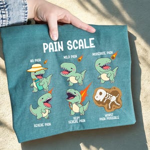 May include: Teal graphic tee featuring a "Pain Scale" with cartoon dinosaurs illustrating levels of pain from "No Pain" to "Worst Pain Possible." The text is in white, and the dinosaurs are green with various expressions.