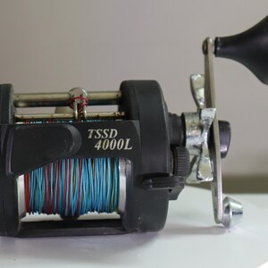 May include: A black TSSD 4000L fishing reel featuring a silver metal handle with a black knob, a star drag adjustment wheel, and a spool wound with bright blue and red fishing line. The reel body is constructed from matte black material.