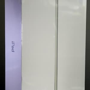 May include: A white rectangular iPad product box standing vertically. The front surface is smooth and glossy, featuring a thin, vertical, light gray line design. The word iPad is printed in small, dark gray text on the side of the box.