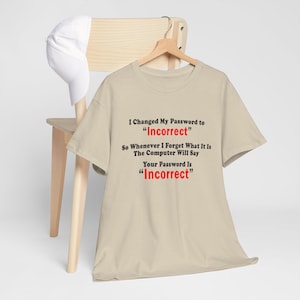 Könnte beinhalten: Beiges T-Shirt mit dem Text "I Changed My Password to 'Incorrect'" in Schwarz und Rot. Das Shirt hängt an einem Holzkleiderbügel, mit einer weißen Baseballkappe auf einem Holzstuhl.