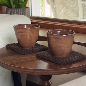 May include: Two brown ceramic cups with a smooth, glossy finish, each resting on a square, dark brown textured coaster. The cups have a simple, handleless design with a slightly tapered shape, showcasing a consistent earthy tone throughout.