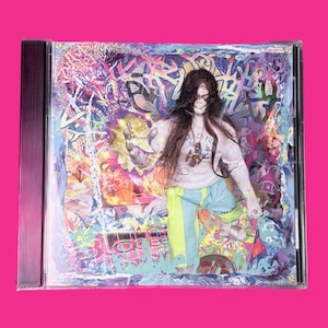 May include: A CD case featuring a colorful, abstract design with a figure in the center. The figure is wearing a light-colored sweatshirt, blue pants with yellow stripes, and long dark hair. The background is a mix of vibrant colors.