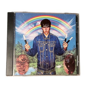 May include: CD case featuring a digitally altered image of a person in a denim jacket holding two black and white objects, with a rainbow and two dogs in the background. The image has a surreal, artistic style.
