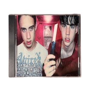 May include: A CD case featuring two young men. One wears a white t-shirt with blue text, the other a red shirt and holds a knife. The background is a red-toned room with a window.
