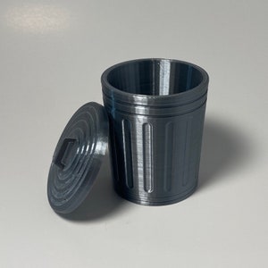 May include: A small, gray, plastic trash can with a lid. The trash can has a ribbed texture and a slightly rounded shape.