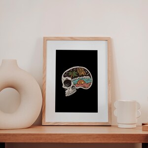 May include: A framed art print featuring a skull with a colorful beach scene inside. The words "GOOD TIMES" are written above the beach scene. The print is displayed on a wooden table with a vase, mug, and hourglass.
