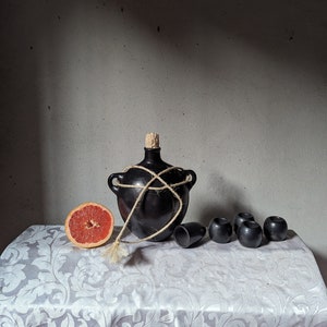 May include: A black ceramic bottle with a cork stopper and a rope tied around it, a half grapefruit, and six small black ceramic cups on a white tablecloth with a floral pattern.