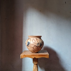 Puede incluir: Un jarrón de cerámica marrón con un diseño decorativo negro y marrón se encuentra sobre una mesa de madera con una sola pata de pedestal.