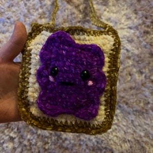 Könnte beinhalten: Eine gehäkelte, violette, mit Gelee gefüllte Toast-förmige Tasche mit einem goldenen Kettenriemen. Die Tasche hat ein süßes Comic-Gesicht mit schwarzen Augen und einem rosafarbenen Lächeln.