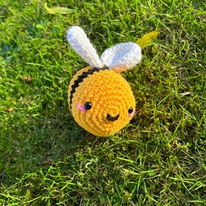 May include: A crocheted bee toy with yellow and black stripes, white wings, and a smiling face with pink cheeks. The bee is sitting on a bed of green grass.