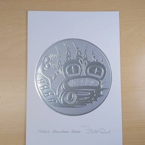Peut inclure: Une estampe argentée en relief représentant un tambour à tête de taureau Haida avec la signature de l'artiste "Bill Reid" sous l'image.