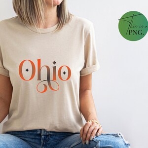 Puede incluir: Una camiseta beige con la palabra "Ohio" impresa en letras naranja y negras con un adorno decorativo.