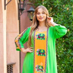 May include: A green and orange maxi dress with embroidered elephants. The dress has a V-neckline and long sleeves with cutouts at the shoulders. The dress is decorated with a colorful trim.