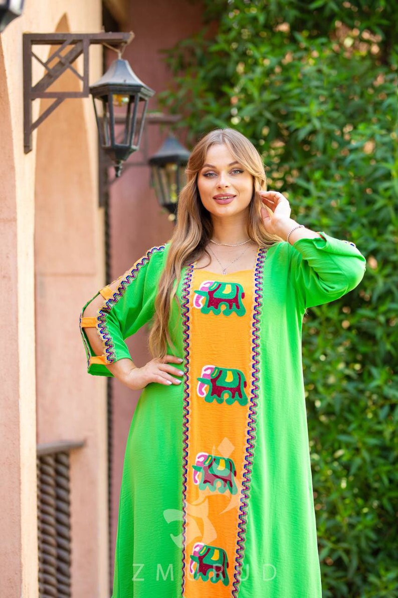 May include: A green and orange maxi dress with embroidered elephants. The dress has a V-neckline and long sleeves with cutouts at the shoulders. The dress is decorated with a colorful trim.