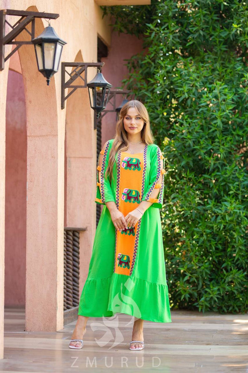 May include: A woman wearing a green and orange maxi dress with embroidered elephants. The dress has a ruffled hem and three-quarter sleeves with cutouts. The woman is standing in front of a brick wall with a green bush in the background.