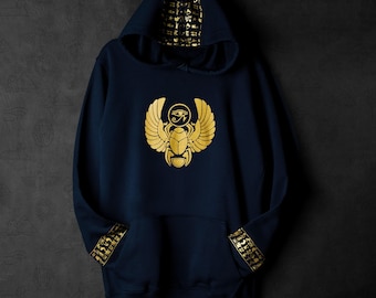 Pode incluir: Uma camisola de capuz azul marinho com detalhes dourados. A frente da camisola apresenta um desenho de escaravelho dourado com asas e o Olho de Hórus. O capuz e as mangas da camisola são forrados com hieróglifos dourados.