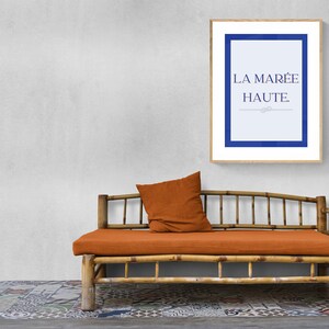 May include: A framed print with a blue border and white background. The text "LA MAREE HAUTE" is printed in blue on the print. The print is hanging on a white wall above a brown bamboo couch with an orange cushion.