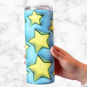 May include: A stainless steel tumbler with a light blue background and yellow stars. The stars have a 3D effect. The tumbler is cylindrical and has a clear lid. The tumbler is held by a person's hand.
