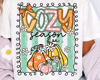 Puede incluir: Camiseta blanca con un gráfico colorido de "COZY season". El diseño presenta calabazas, flores y un lazo, todo dentro de un borde decorativo. La camiseta es una prenda cómoda e informal.