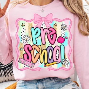 May include: Pink sweatshirt with a colorful "Pre School" graphic. The design includes a pink bow, stars, flowers, a ruler, a pencil, and an apple. The background has a gold glitter stripe pattern.