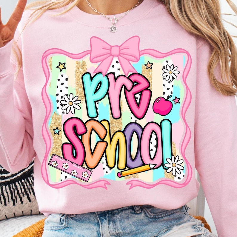 May include: Pink sweatshirt with a colorful "Pre School" graphic. The design includes a pink bow, stars, flowers, a ruler, a pencil, and an apple. The background has a gold glitter stripe pattern.