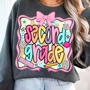 May include: Dark gray sweatshirt featuring a colorful graphic that reads "Second Grade." The design includes a pink bow, daisies, stars, a ruler, a pencil, and an apple. The text is in a variety of bright colors.