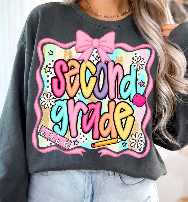 May include: Dark gray sweatshirt featuring a colorful graphic that reads "Second Grade." The design includes a pink bow, daisies, stars, a ruler, a pencil, and an apple. The text is in a variety of bright colors.