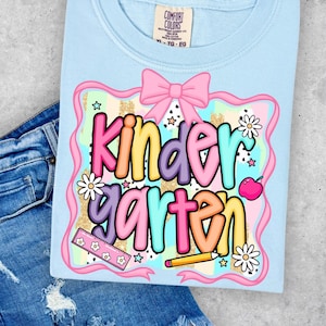 May include: A light blue t-shirt with the word "Kindergarten" in colorful bubble letters. The design includes a pink bow, flowers, a ruler, a pencil, and an apple. The shirt is paired with blue denim jeans.