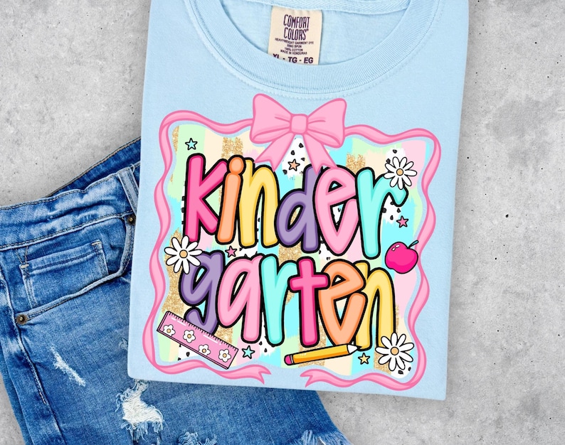 May include: A light blue t-shirt with the word "Kindergarten" in colorful bubble letters. The design includes a pink bow, flowers, a ruler, a pencil, and an apple. The shirt is paired with blue denim jeans.
