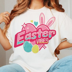 May include: White t-shirt with a pink heart design that says "Easter Vibes" in colorful letters. The heart has bunny ears and two pastel Easter eggs.