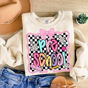 May include: Cream-colored sweatshirt with the word "PRE SCHOOL" in colorful, playful lettering. The design includes a pink bow, daisies, and a checkerboard pattern. The sweatshirt is paired with blue denim shorts and sandals.