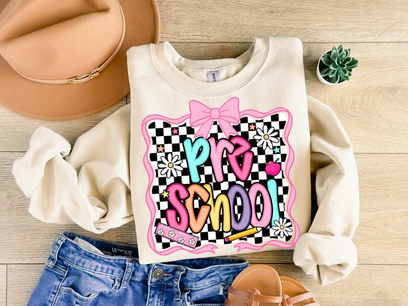 May include: Cream-colored sweatshirt with the word "PRE SCHOOL" in colorful, playful lettering. The design includes a pink bow, daisies, and a checkerboard pattern. The sweatshirt is paired with blue denim shorts and sandals.