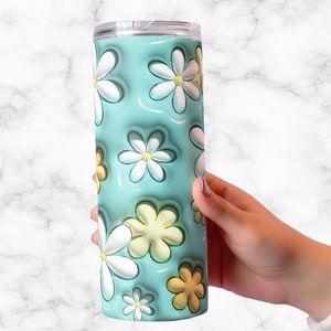 May include: A floral-themed tumbler with a light blue background and white and yellow daisy designs. The tumbler is cylindrical and has a clear lid. The design features a 3D effect, giving the flowers a raised appearance.