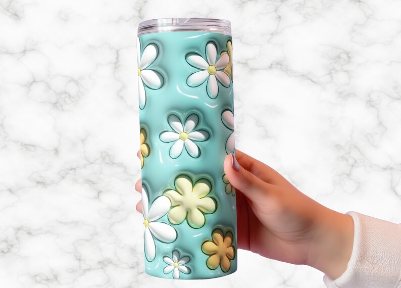 May include: A floral-themed tumbler with a light blue background and white and yellow daisy designs. The tumbler is cylindrical and has a clear lid. The design features a 3D effect, giving the flowers a raised appearance.