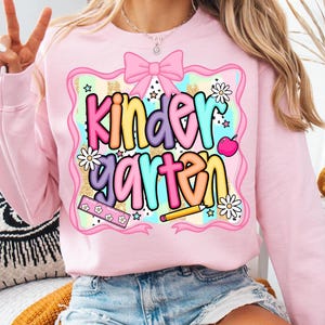 May include: Pink sweatshirt with a colorful "Kindergarten" graphic. The design includes a pink bow, daisies, stars, a ruler, a pencil, and an apple. The text is in multiple colors, including yellow, purple, and blue.