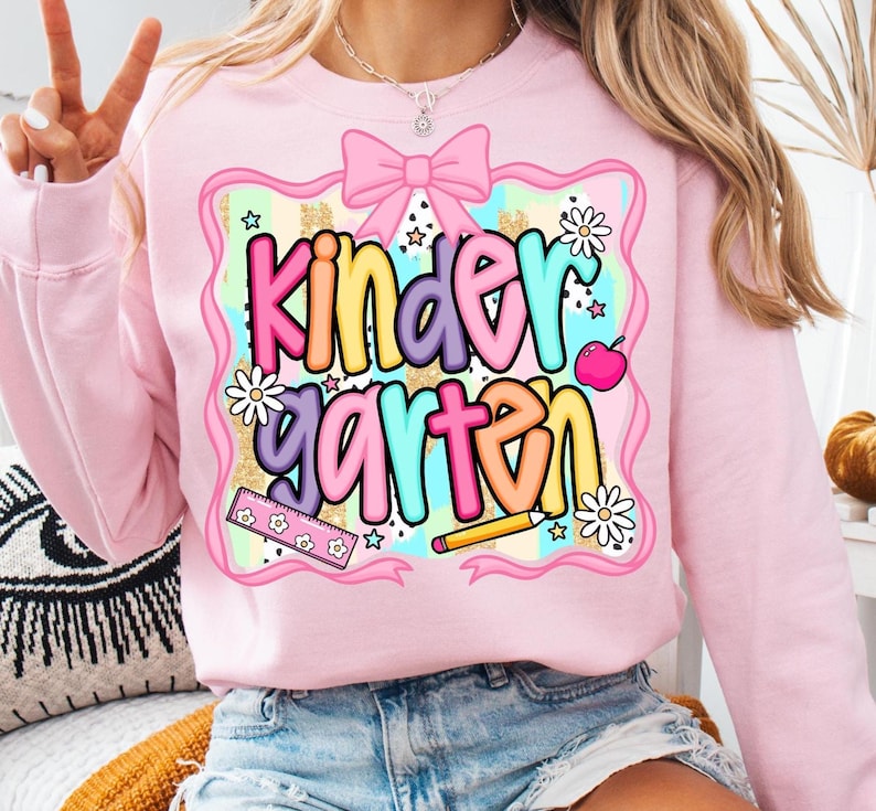 May include: Pink sweatshirt with a colorful "Kindergarten" graphic. The design includes a pink bow, daisies, stars, a ruler, a pencil, and an apple. The text is in multiple colors, including yellow, purple, and blue.