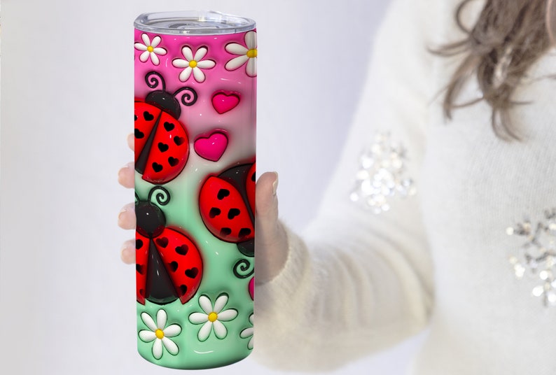 K&ouml;nnte beinhalten: Ein Edelstahlbecher mit einem rosa-gr&uuml;nen Ombre-Design mit Marienk&auml;fern, Herzen und G&auml;nsebl&uuml;mchen.